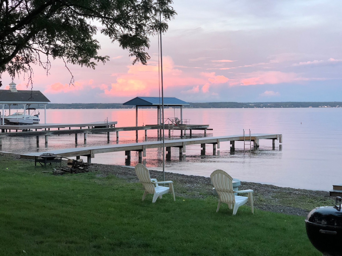 A serene lakeside view is presented, featuring two white Adirondack chairs on the grassy shore. A wooden dock extends into the calm water, under a sky showcasing soft pink and purple hues at sunset. A gazebo is visible in the distance, adding to the peaceful atmosphere.