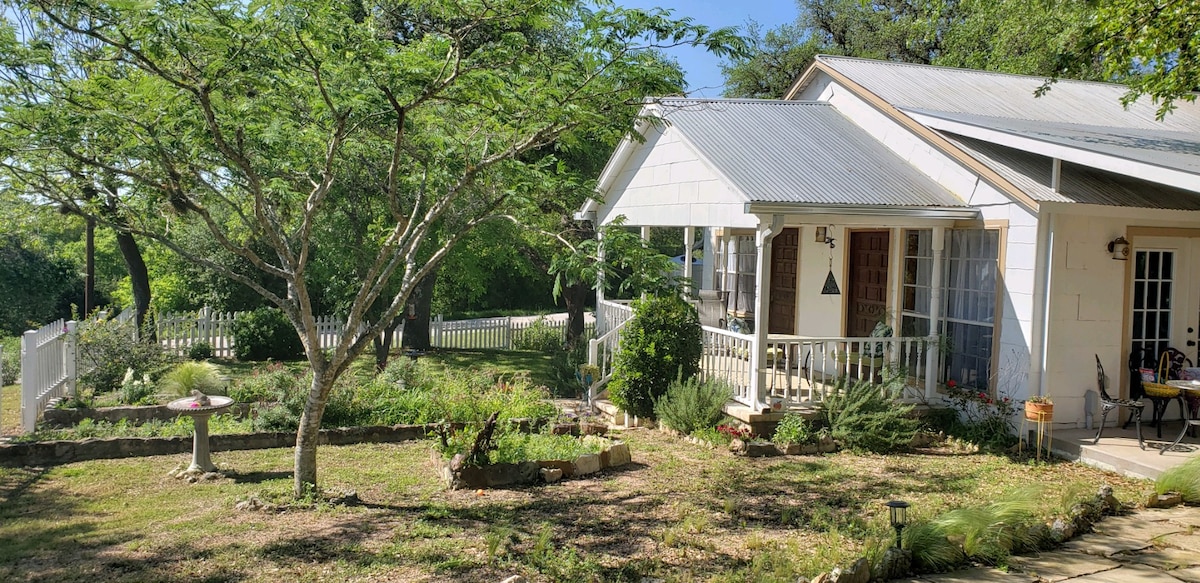 The exterior of the house features a charming porch with a white railing, surrounded by lush green landscaping. Multiple trees provide shade to the outdoor area, which is enhanced by a landscaped garden and stone pathways leading up to the entrance.