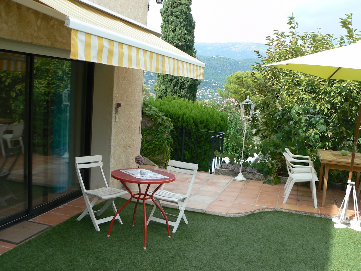 A private terrace features a circular red table surrounded by four white chairs, with an umbrella providing shade. The terrace is adorned with artificial grass and offers a view of the surrounding greenery and hills, complemented by a striped awning.