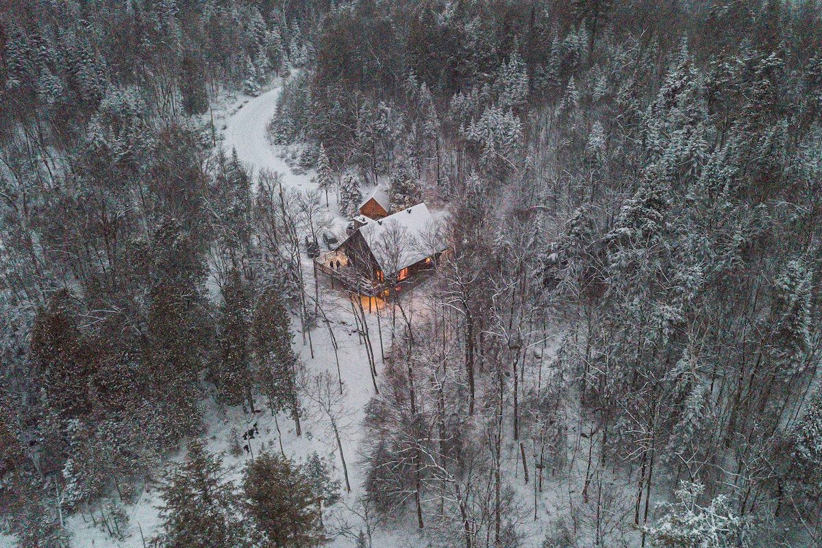 An aerial view captures a chalet nestled among snow-covered trees, surrounded by a winter landscape. Warm light emits from the windows, contrasting with the white snow and creating a cozy appearance. The winding road is visible, leading to the entrance of the property.