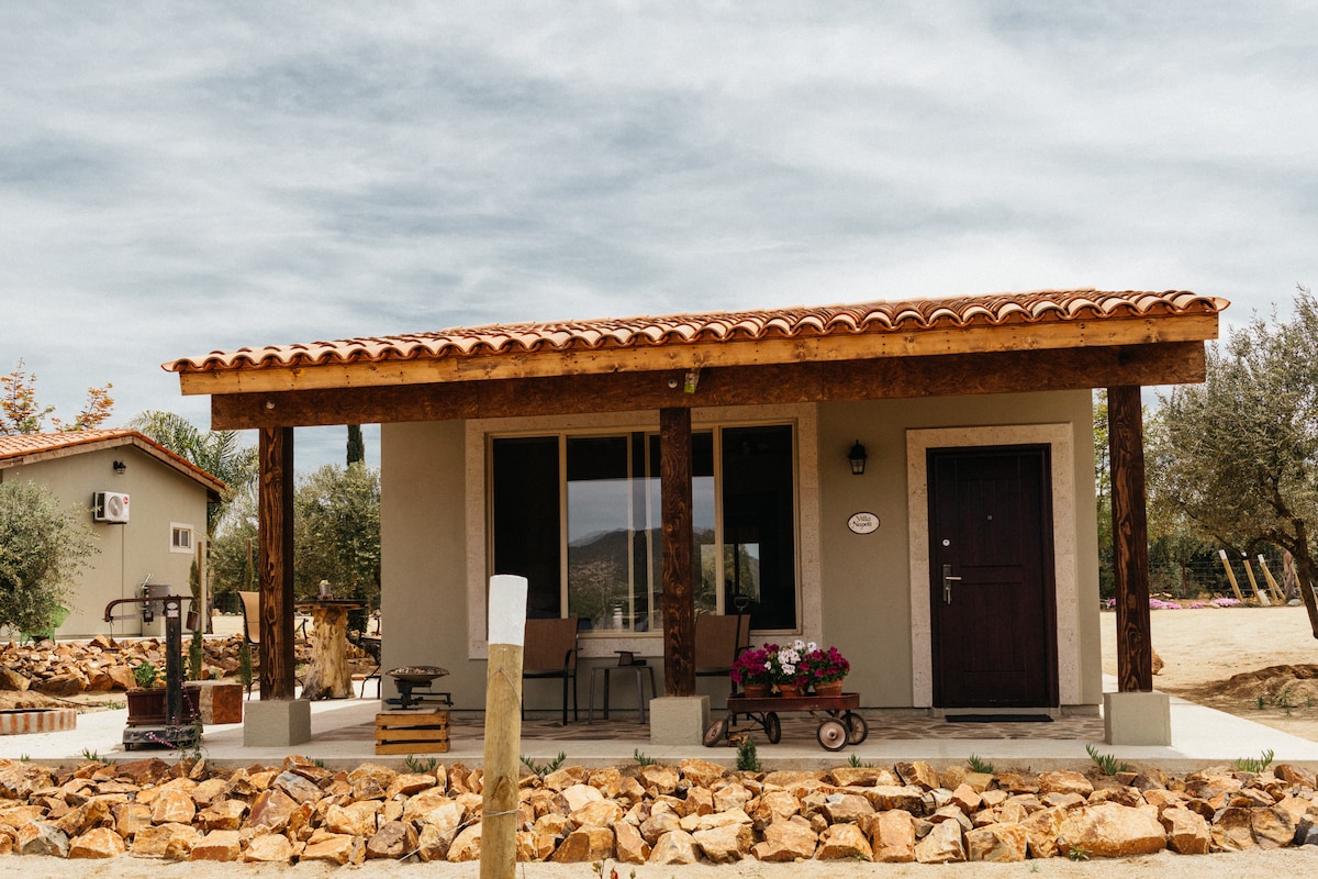 The exterior of the villa is depicted, featuring a covered porch supported by wooden beams. Two chairs and a small table are arranged on the porch, complemented by decorative flowers. The entrance door is framed by a warm, natural stone pathway, surrounded by greenery.
