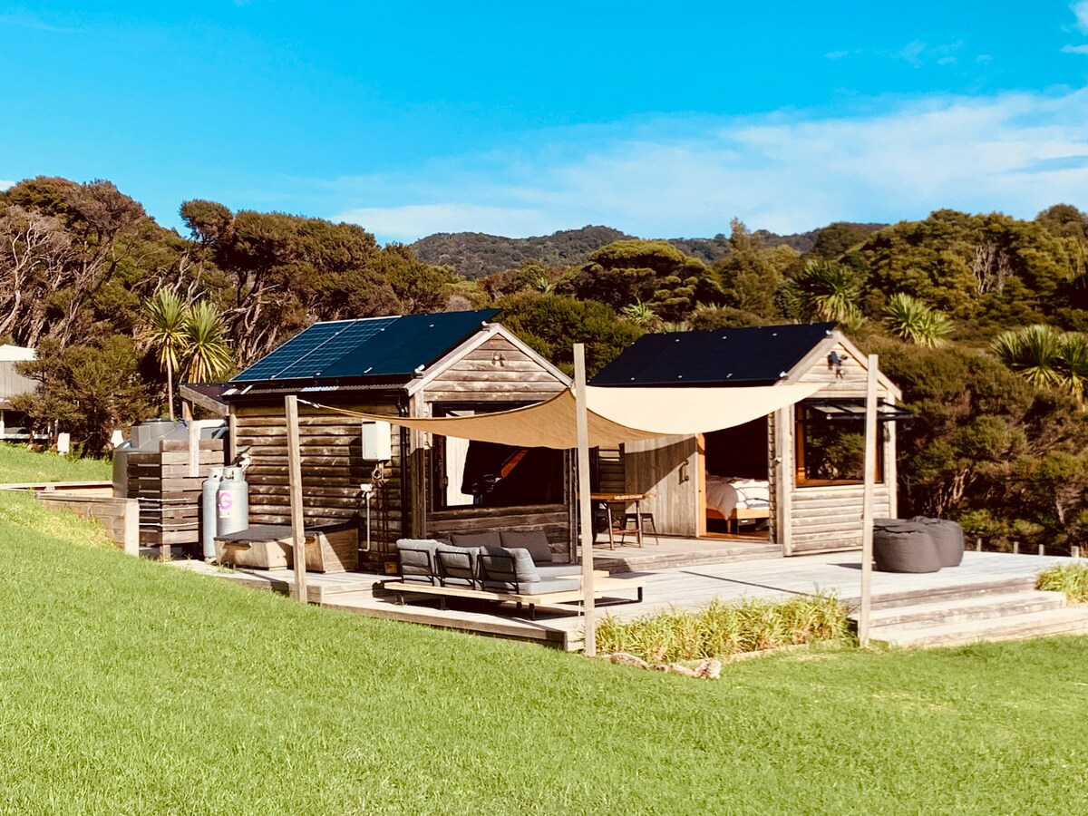 Two rustic cabins are situated on a grassy area, connected by a spacious wooden deck. Solar panels sit atop the structures. A shaded seating area with outdoor furniture is visible, offering a comfortable space to relax. Lush bush surrounds the cabins, creating a natural setting.