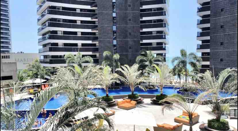 A vibrant outdoor area is showcased, featuring a large swimming pool surrounded by lush palm trees and lounge chairs. The contemporary architecture of the surrounding buildings is visible, with a sunny sky overhead, creating an appealing environment for relaxation.