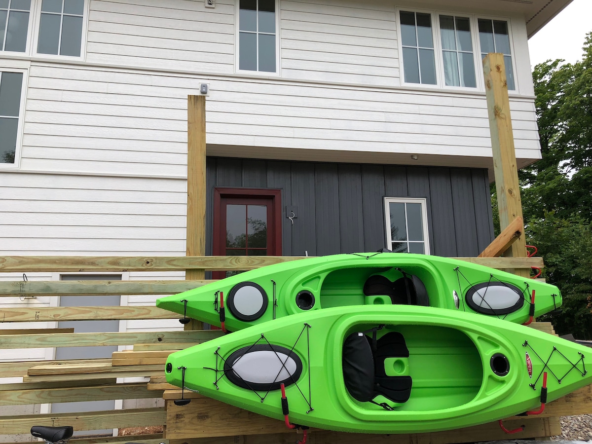 Two bright green kayaks are positioned on a wooden rack outside the modern cottage, which features a clean, white exterior and large windows. The entrance is adorned with a dark door, enhancing the welcoming appearance of the property.
