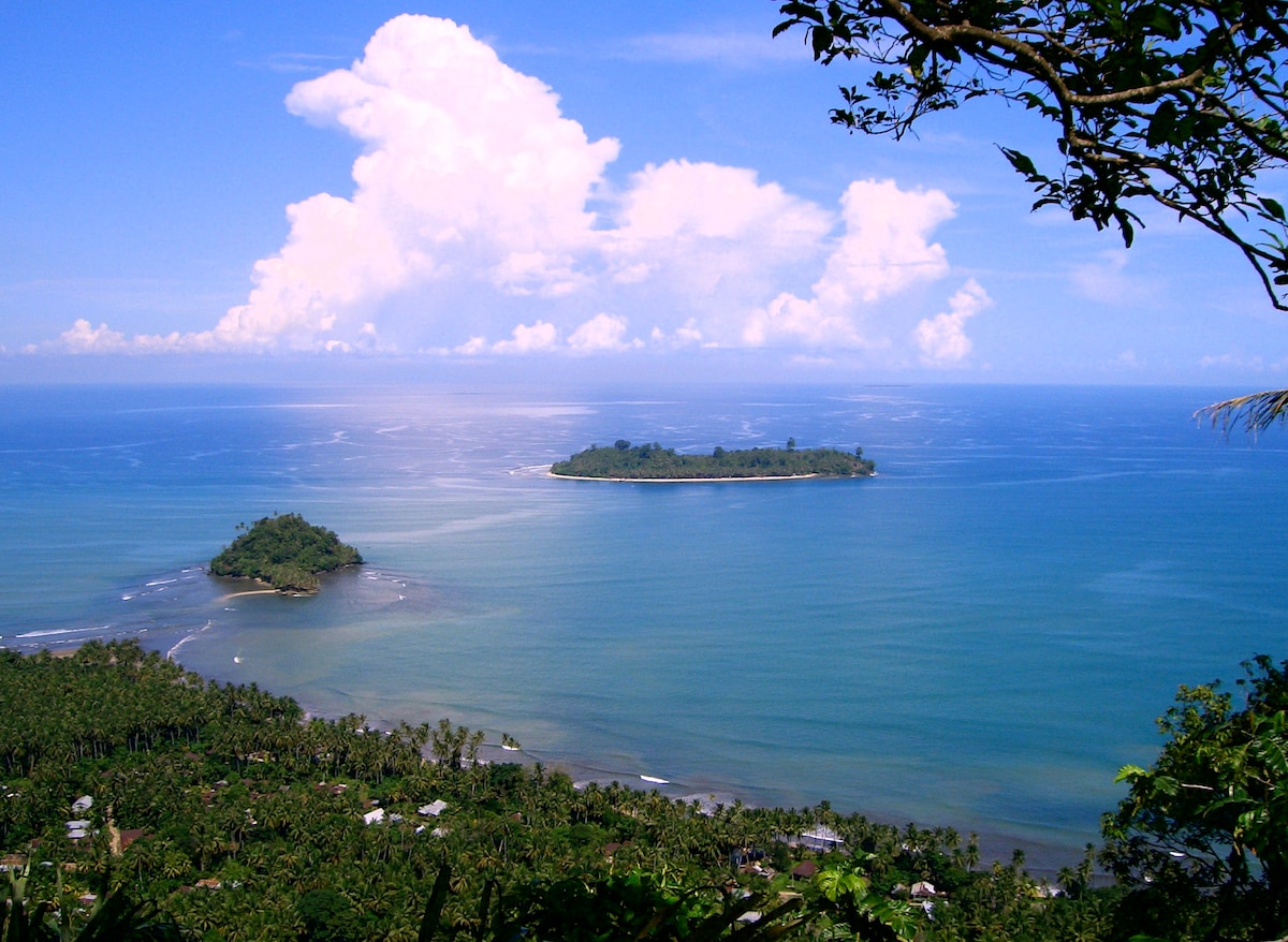A panoramic view of the coastline is captured, showcasing the expansive blue ocean with gentle waves. Islands are visible in the distance, surrounded by lush greenery. White clouds are scattered across a bright blue sky, reflecting a serene and tranquil atmosphere.