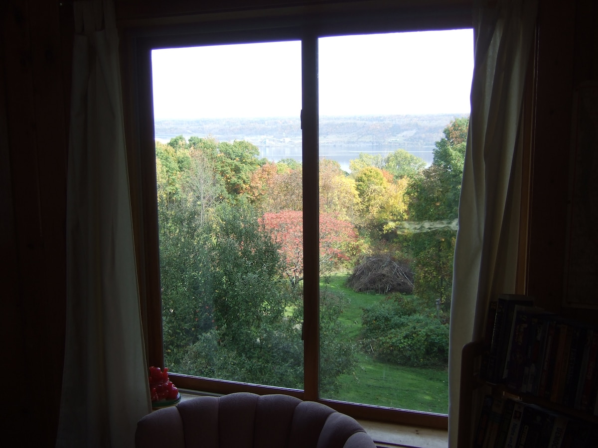 A large window frames a vibrant view of rolling hills and colorful foliage in the foreground. The expansive landscape extends toward a lake in the distance, enhancing the serene ambiance of the rural setting.