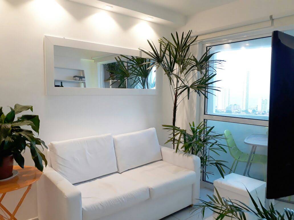 A bright living area features a white sofa across from a large mirror, enhancing the room's openness. Potted plants are positioned around the space, adding a touch of greenery. Natural light filters in through a large window, offering a view of the surrounding cityscape.
