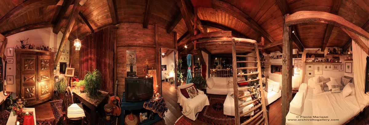 A panoramic view of a warmly lit living area features wooden beams and rustic decor. A spacious seating arrangement is complemented by a cozy blanket draped over a couch. A TV unit and decorative elements add character to the space, while a staircase leads to a loft area.