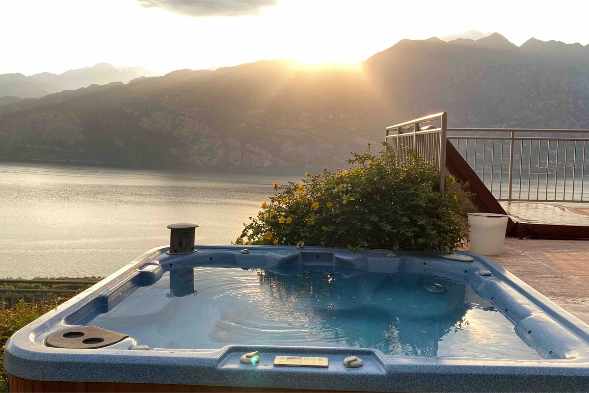 A hot tub is positioned on a terrace, offering a serene view of Lake Garda. Soft sunlight reflects off the water's surface, while the surrounding greenery adds a natural element. The backdrop features majestic mountains, enhancing the tranquil setting.