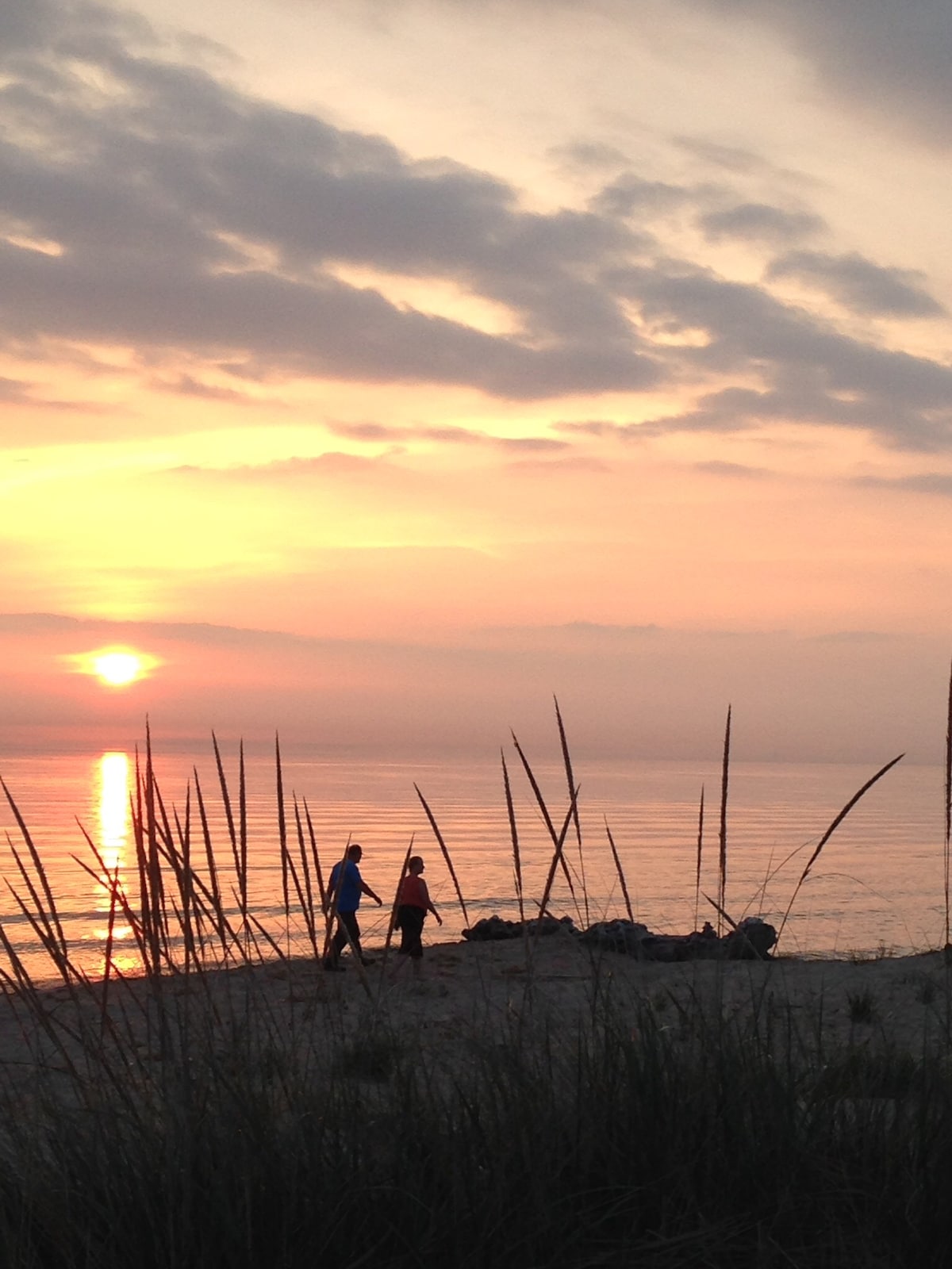 A serene beach scene captures two figures walking along the shore, silhouetted against a colorful sunset. Soft sand is framed by tall grasses, with gentle waves lapping in the background. The sky is painted in warm hues, reflecting the tranquil atmosphere of the setting.