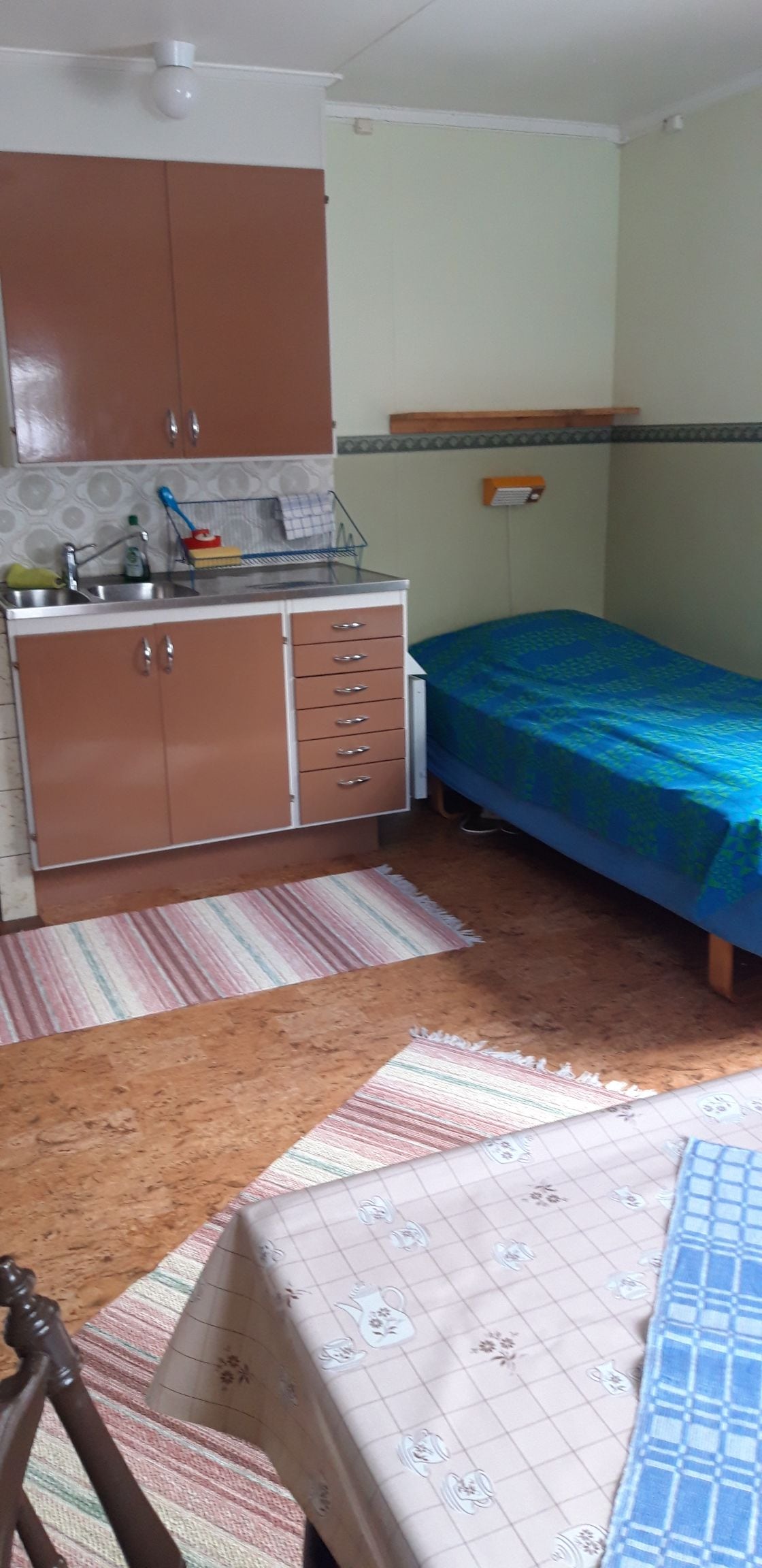 The image shows a compact accommodation space featuring a single bed with a blue patterned cover. A kitchenette is situated along one wall, equipped with cabinets and counter space. Two distinct area rugs are present on the cork floor, adding texture and warmth to the room.