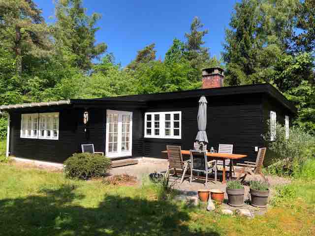 A charming black wooden house is surrounded by lush greenery, featuring large windows that allow ample natural light. An outdoor dining area with a table and chairs is visible, complemented by potted plants and a patio umbrella, creating an inviting space for relaxation.