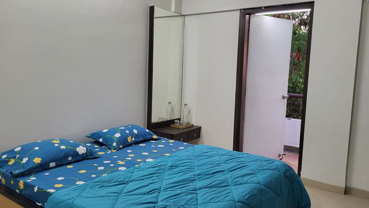 A well-lit bedroom featuring a spacious bed with a blue floral bedspread. A mirror wardrobe reflects the room's light, and an open door leads to a small balcony area with greenery visible outside.