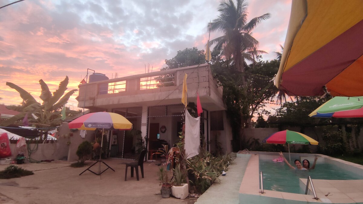 A vibrant outdoor space is defined by colorful umbrellas above a concrete patio. A two-story building stands in the background, framed by tropical greenery. The inviting pool reflects the warm hues of the sunset, creating a serene atmosphere for relaxation.