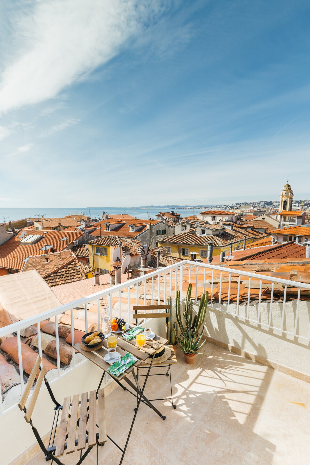 A rooftop terrace is shown with a small table set for breakfast, featuring drinks and pastries. Scenic views of terracotta rooftops and the sea are visible in the distance, complemented by a potted plant. Bright blue skies and sunlight create a cheerful ambiance.