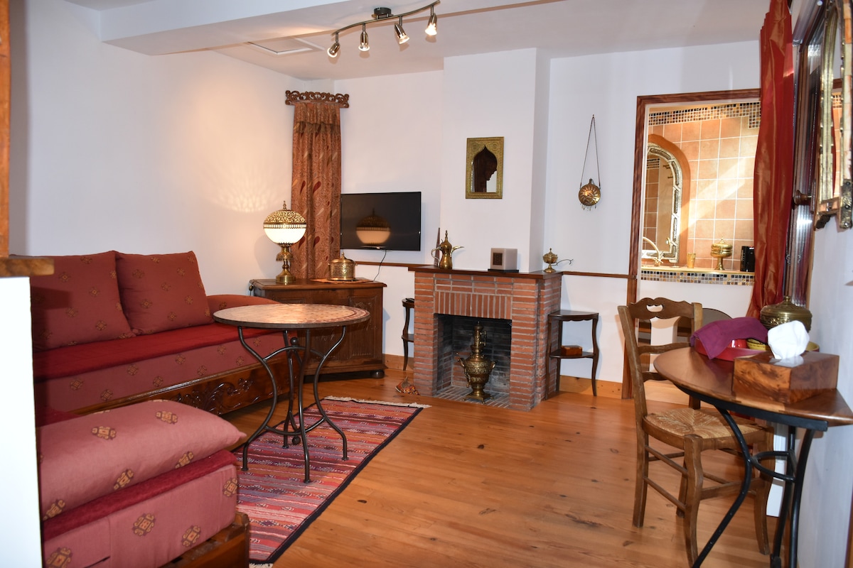 A cozy lounge area features a comfortable red sofa and a decorative fireplace. A flat-screen TV is mounted on the wall, complemented by various decorative elements. A round table with chairs is positioned nearby, and warm wood flooring enhances the inviting space.