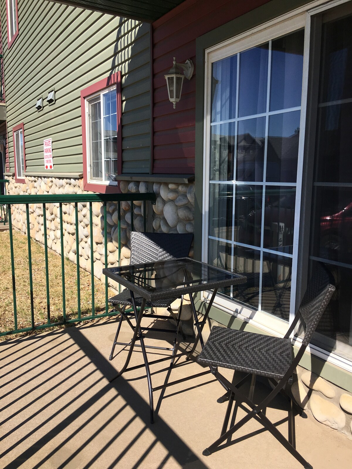 A small outdoor patio is presented, featuring a glass-top table accompanied by two black wicker chairs. Sunlight casts shadows through the railing, highlighting the stone facade and the nearby green and red siding of the building. A window reflects the outside scenery.
