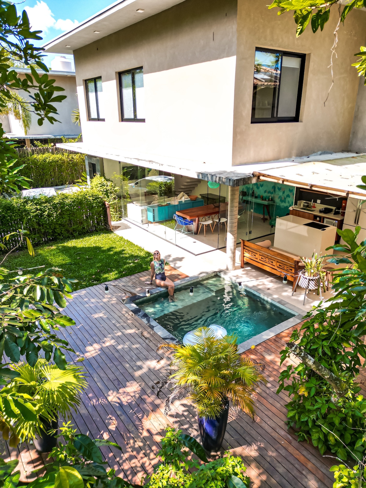 A spacious outdoor area features a private swimming pool surrounded by wooden decking and lush greenery. The two-story house, visible in the background, has large windows allowing natural light to fill the interior. The patio is furnished for relaxation and dining.