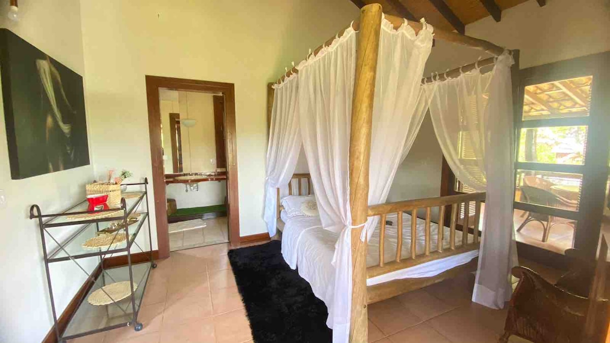 A rustic sleeping area features a wooden four-poster bed draped with white curtains. A cozy area rug lies beneath the bed, and a small metal cart holds various items. Large windows provide natural light and open to the outdoors.