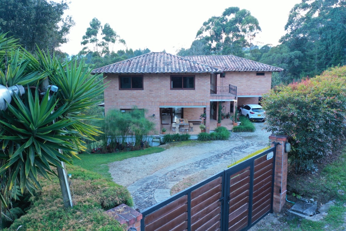 The exterior of a two-story brick house is framed by lush greenery and vibrant plants. A stone pathway leads to the entrance, while a spacious lawn surrounds the property. An electric gate is visible in the foreground, providing an impression of privacy and security.