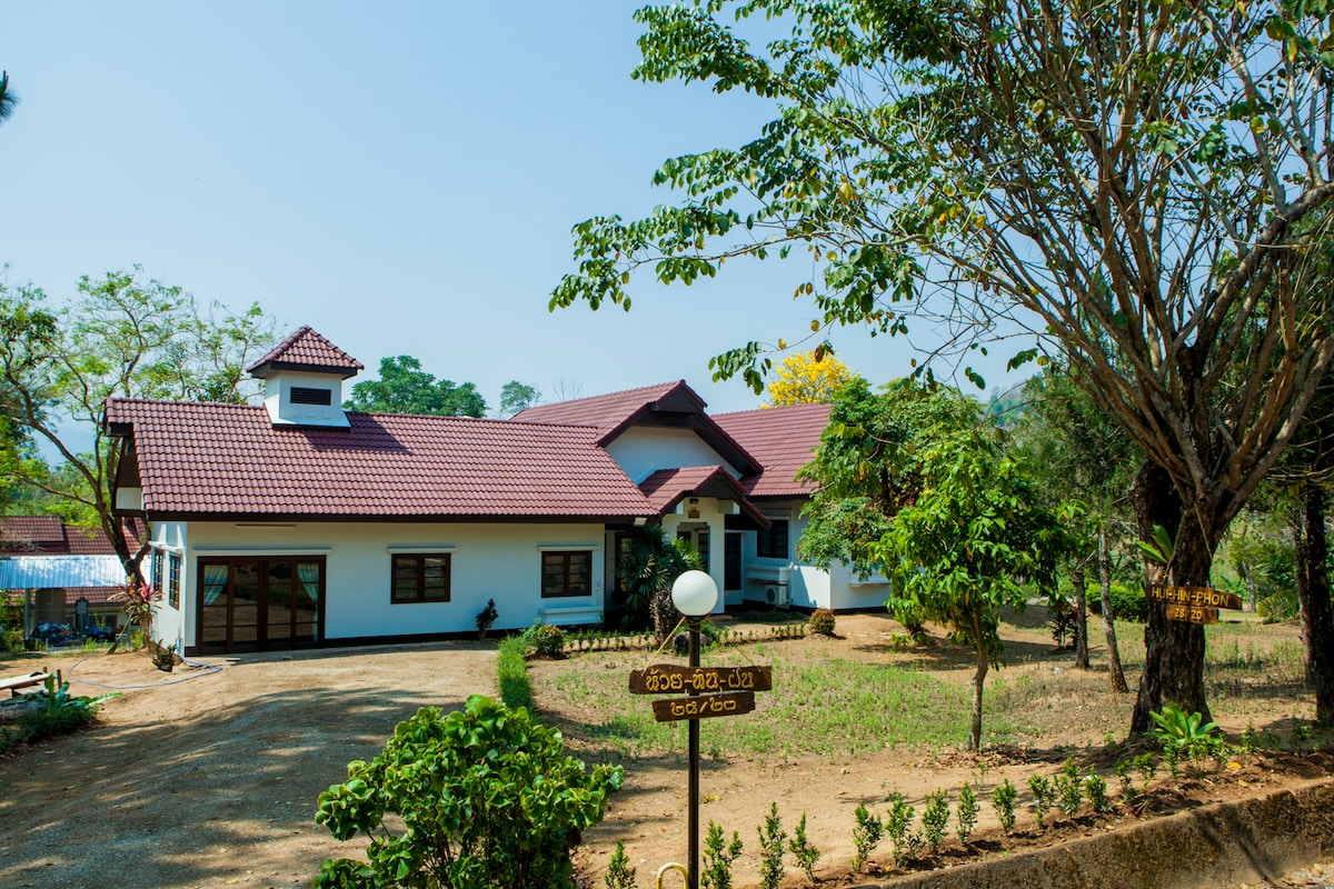 A private house is presented amid a tranquil landscape, featuring a sloping roof and a welcoming entrance. Surrounding trees and neatly trimmed shrubs enhance the outdoor area. A gravel driveway leads up to the home, providing easy access in a peaceful setting.