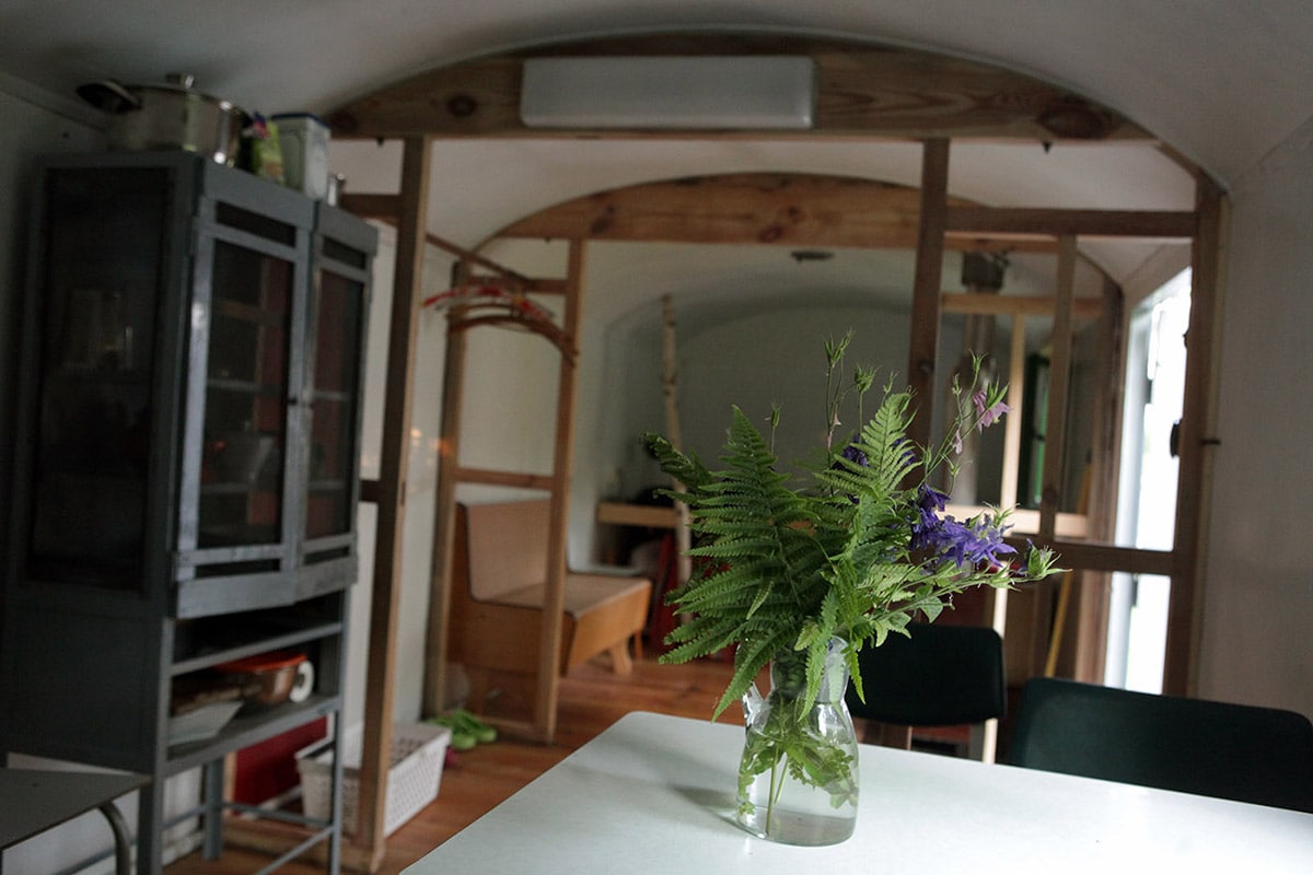 An interior view of a restored trailer features a small dining table with a vase of fresh flowers. Wooden framing is visible throughout the space. A cozy sleeping area is set in the background, with natural light filtering through the windows.