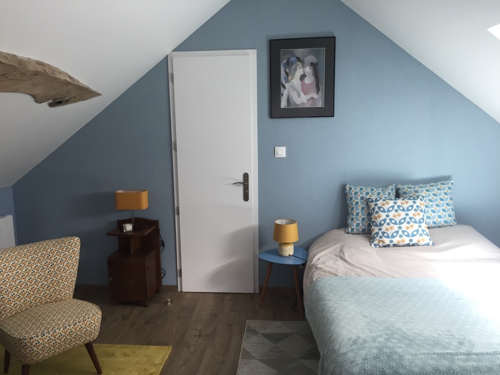 A cozy room is featured with a sloped ceiling and painted blue walls. A spacious bed is adorned with colorful patterned pillows. A small wooden nightstand and a blue table with a lampshade add functionality, alongside a patterned chair positioned nearby.