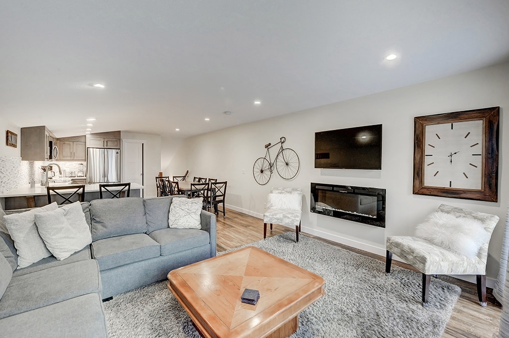 A spacious living area features a sectional sofa in light grey, accented by two plush white chairs. A wooden coffee table is centered on a soft rug. A large flat-screen TV is mounted on the wall, with a modern clock nearby. The open kitchen and dining area are visible in the background.