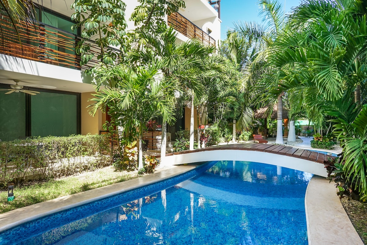 A serene pool area is surrounded by lush greenery, with tall palm trees and vibrant plants framing the water. The pool features a gentle curve and a wooden deck, creating a peaceful outdoor retreat. Sunlight reflects off the clear water, enhancing the relaxing ambiance.