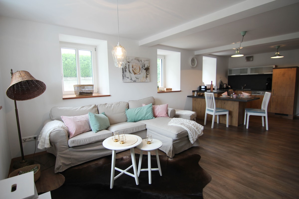 A bright living area features a light-colored sofa adorned with pastel cushions, complemented by two small white tables. A wood dining table is visible in the background, accompanied by white chairs. Natural light enters through two windows, illuminating the warm wooden floor.