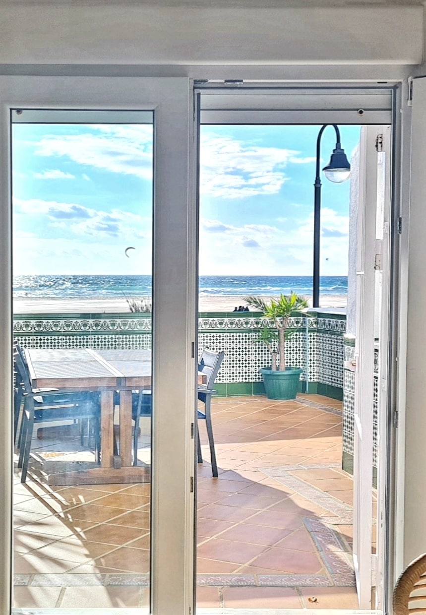 A bright terrace door opens to reveal a view of the sea. A large dining table is situated on the terrace, surrounded by seating. A potted plant adds a touch of greenery, while the sky reflects various shades of blue with scattered clouds.