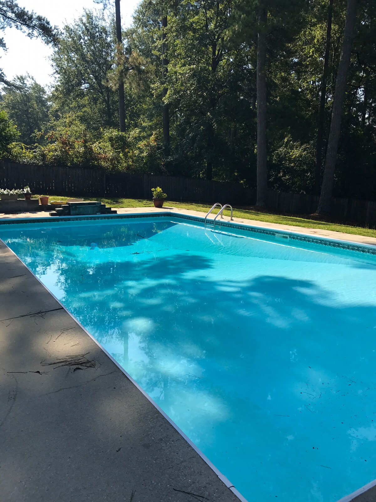 An inground pool is surrounded by a concrete deck, featuring a clear blue water surface. Lush greenery and trees provide shade along the perimeter, with potted plants located on the edge of the deck. Steps lead into the shallow end of the pool.