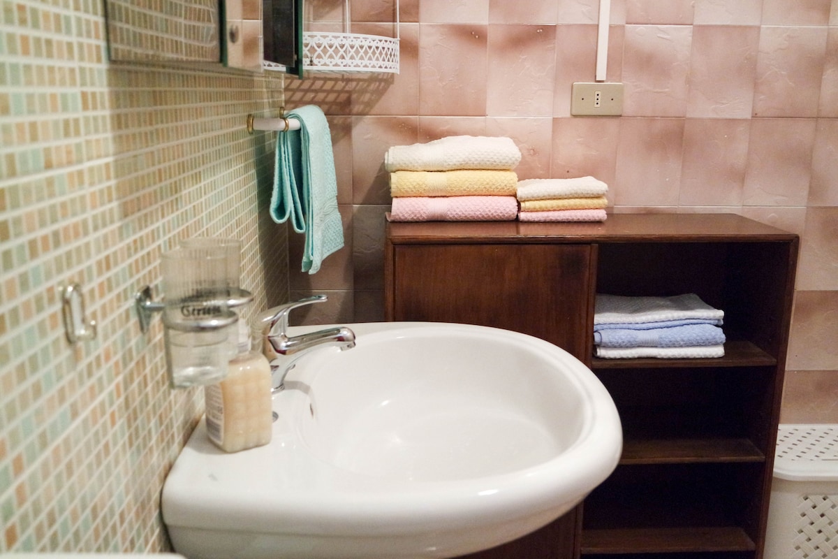 The bathroom features a spacious sink with a modern faucet, accompanied by a wooden shelf displaying neatly folded towels in soft pastel colors. A mosaic-tiled wall provides a textured backdrop, complementing the overall arrangement. A small soap dispenser and a glass container are visible on the counter.
