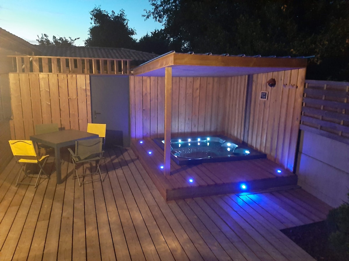 An outdoor area features a wooden deck illuminated by soft blue lights, encompassing a private hot tub. A shaded seating area with a table and four chairs is positioned nearby, surrounded by wooden paneling for added privacy.