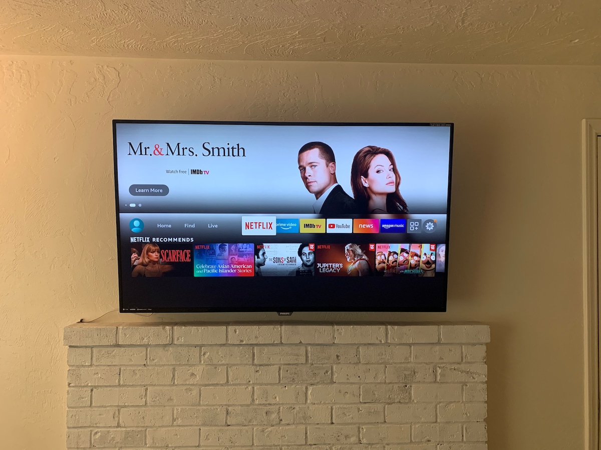 A 55-inch 4K television is mounted above a white brick fireplace, displaying the menu screen for popular streaming services. The layout shows various movie options and recommendations, providing guests with entertainment choices during their stay.