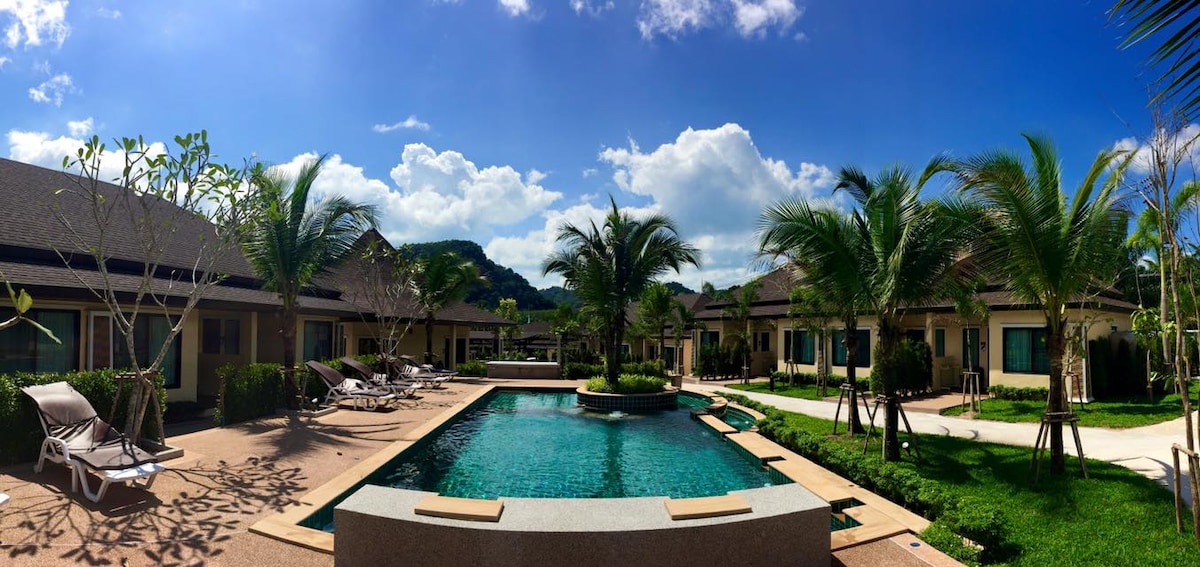 A panoramic view of the hotel grounds reveals a shared swimming pool surrounded by lounge chairs and palm trees. The buildings are designed in a contemporary style, with large windows allowing natural light to enter. Lush greenery enhances the outdoor space under a clear blue sky.