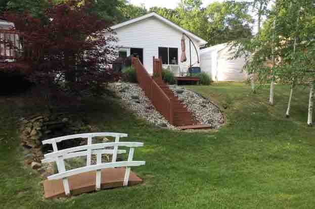 A well-maintained cottage is visible, surrounded by greenery. Steps lead up to the entrance, framed by decorative stones and a wooden railing. A small white bridge is positioned in the foreground, enhancing the tranquil outdoor setting.