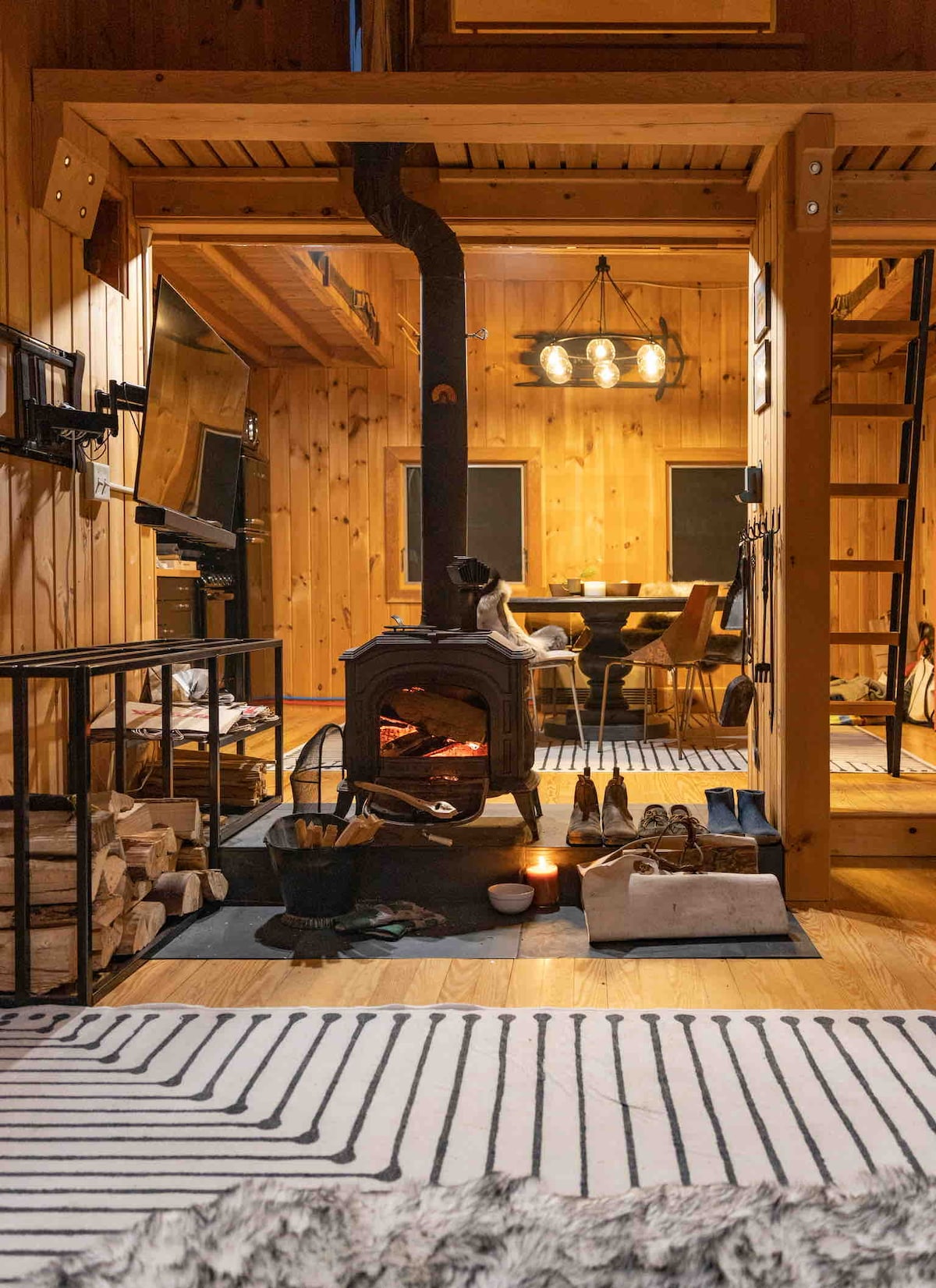A wood stove stands centrally in the living space, providing warmth to the room. Surrounding elements include neatly stacked firewood and rustic decor. A dining area can be seen in the background, illuminated by pendant lighting. The wooden walls enhance the cabin's cozy ambiance.