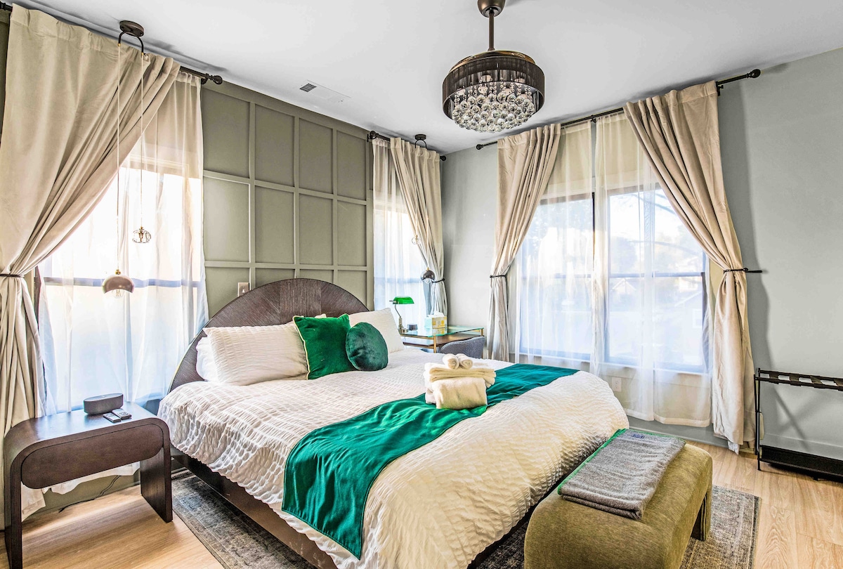 A spacious bedroom features a comfortable bed adorned with crisp white linens and a green accent throw. Natural light floods through two large windows, framed by elegant curtains. A bedside table displays a lamp, and a plush stool sits at the foot of the bed.