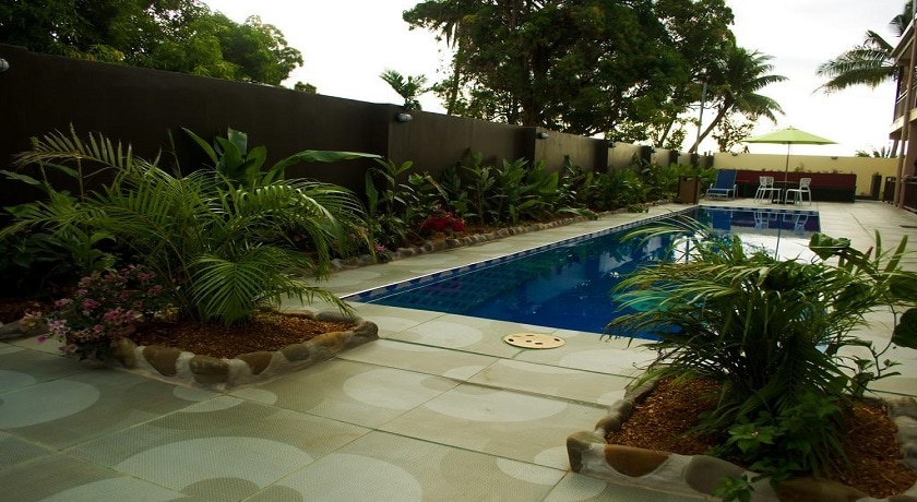 An outdoor swimming pool is framed by tropical plants and landscaped gardens. Sun loungers are positioned nearby under a parasol, providing shaded seating. The pool area features a smooth tile surface, enhancing the natural beauty of the surroundings.