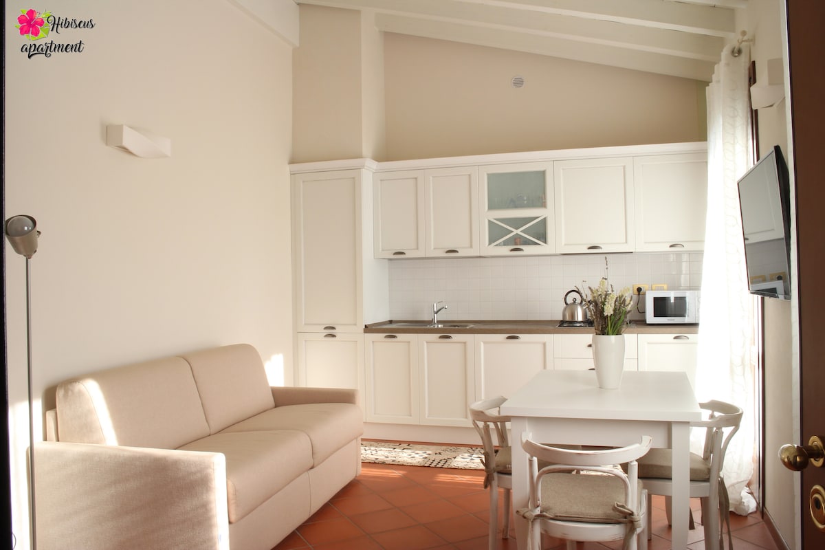 A bright kitchen area features white cabinetry with a tile backsplash. A cozy sofa sits adjacent to a dining table set for four. Natural light floods the space through large windows, complementing the terracotta floor.