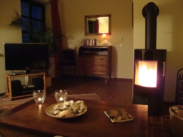 A cozy living area features a black stove providing warmth, with a coffee table displaying decorative shells and candles. A television is positioned nearby, while a wooden dresser and bookshelf are visible in the background. Soft lighting enhances the inviting atmosphere.