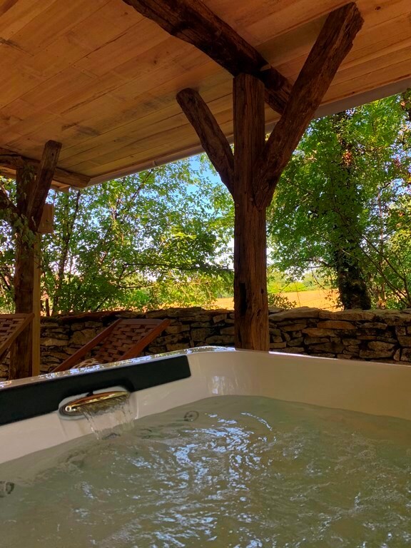 A relaxing private jacuzzi is positioned on a wooden deck area, framed by rustic timber supports. Sunlight filters through lush green trees, casting gentle patterns over the bubbling water. A stone wall enhances the natural setting, creating a serene outdoor space.