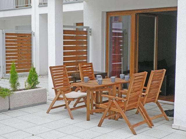 An outdoor terrace is furnished with a wooden dining set, including a table and four chairs. The space is surrounded by low planters and features a backdrop of glass doors leading into the living area. Sunlight illuminates the terrace, enhancing its inviting atmosphere.