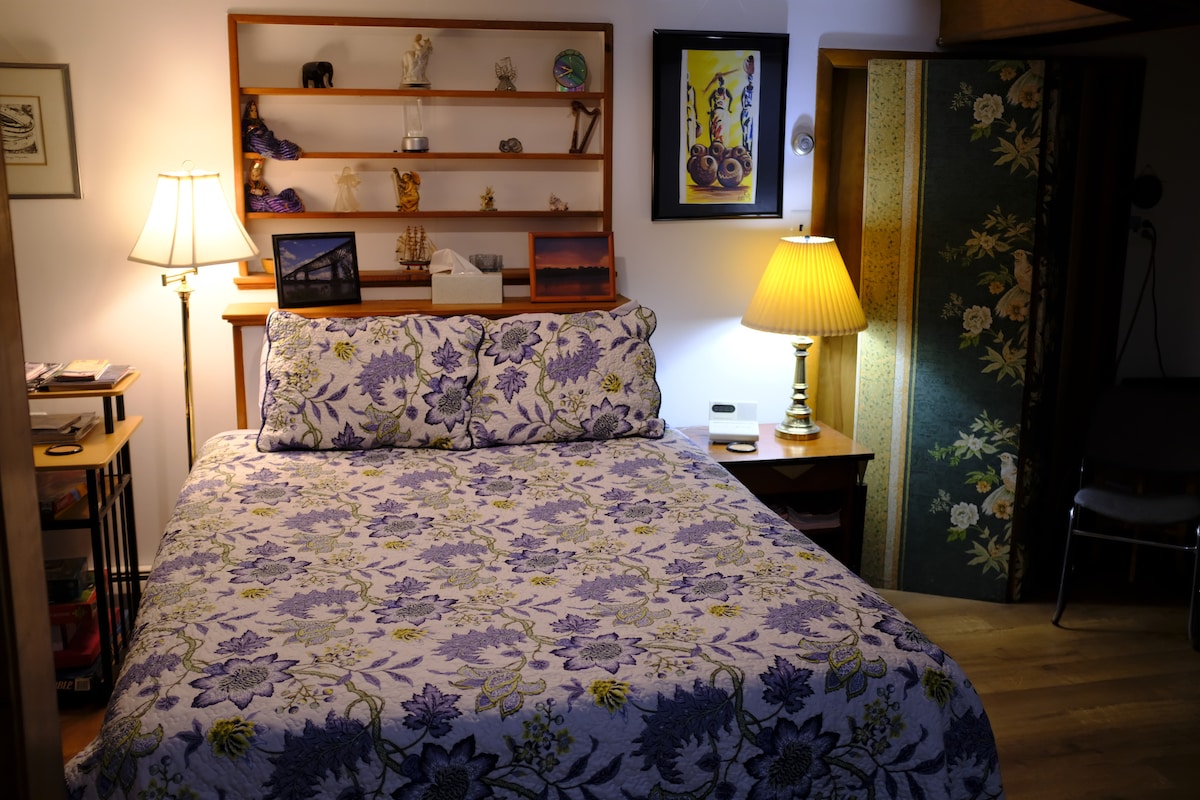 The sleeping area features a queen bed adorned with a floral purple and green comforter. Nightstands with lamps are placed on either side, and additional decor includes shelves with various figurines and artwork on the walls, creating a cozy and inviting space.