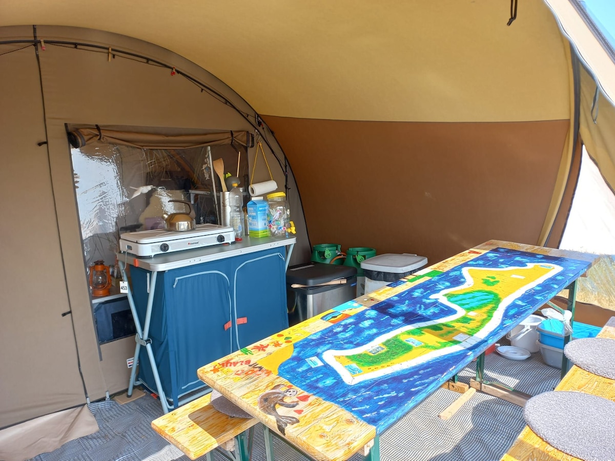 Inside the tent, a kitchen area is visible with a cooking stove and various kitchen supplies arranged on a table. A colorful map design decorates the table's surface, and storage bins are positioned nearby, creating a functional space for meal preparation.