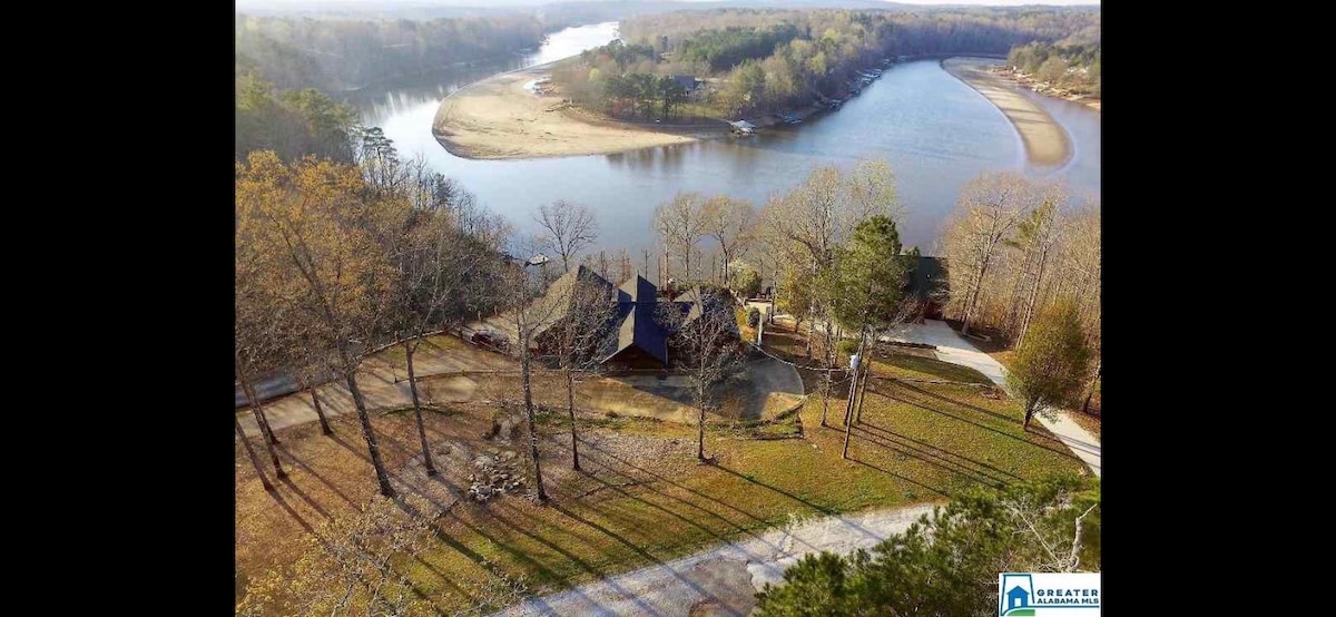 Aerial view of the property reveals a spacious home nestled among trees, with a winding river visible in the background. The landscape showcases a mix of greenery and sandy areas along the water's edge, with paths leading toward the house.