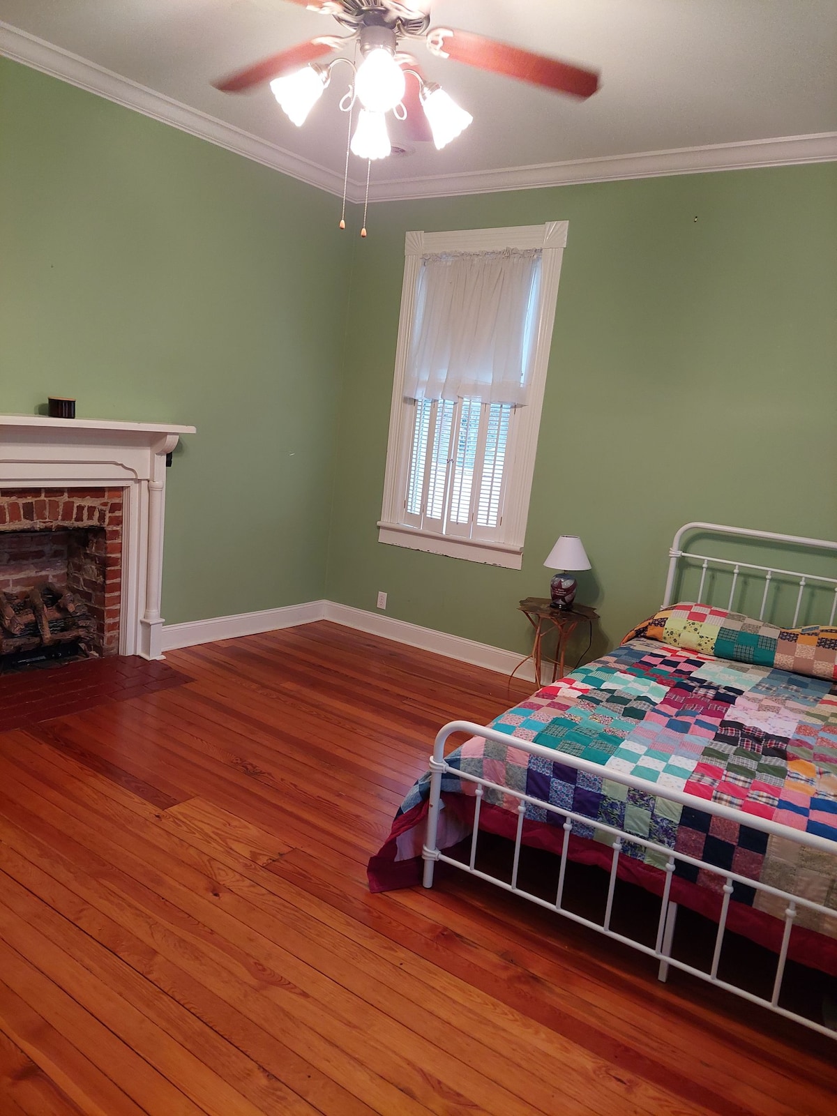 A serene bedroom features a white metal bed adorned with a multicolored patchwork quilt. A brick fireplace adds warmth to the space, while a small nightstand with a lamp is positioned beside the bed. Light filters through a window with sheer curtains, enhancing the calm atmosphere.