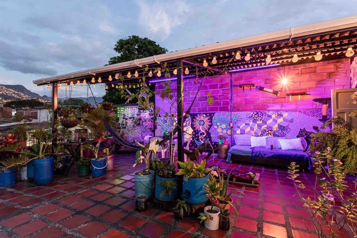 A rooftop terrace is adorned with vibrant plants in colorful pots, and a cozy seating area is highlighted by decorative tiles. String lights provide soft illumination, creating a relaxing atmosphere. A hammock is visible, inviting guests to unwind and enjoy the panoramic views of the surroundings.