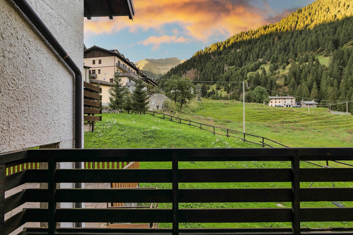 A view from the balcony showcases a lush green landscape, with rolling hills and mountains in the background. A mix of residential buildings is visible, set against the vibrant colors of the sunset sky. The area appears peaceful and inviting.
